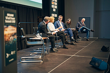 Fünf Personen sitzen auf einer Bühne in einer Podiumsdiskussion. Sie sitzen in einer Reihe auf Stühlen, sprechen miteinander und halten Unterlagen in den Händen. Im Hintergrund sind Sponsorenlogos und eine Leinwand zu sehen.